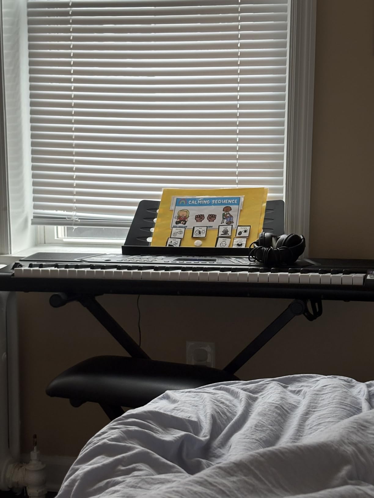 RockJam 61-key keyboard set up on its stand by a bedroom window, headphones resting on the panel and a printed sheet on the music rest