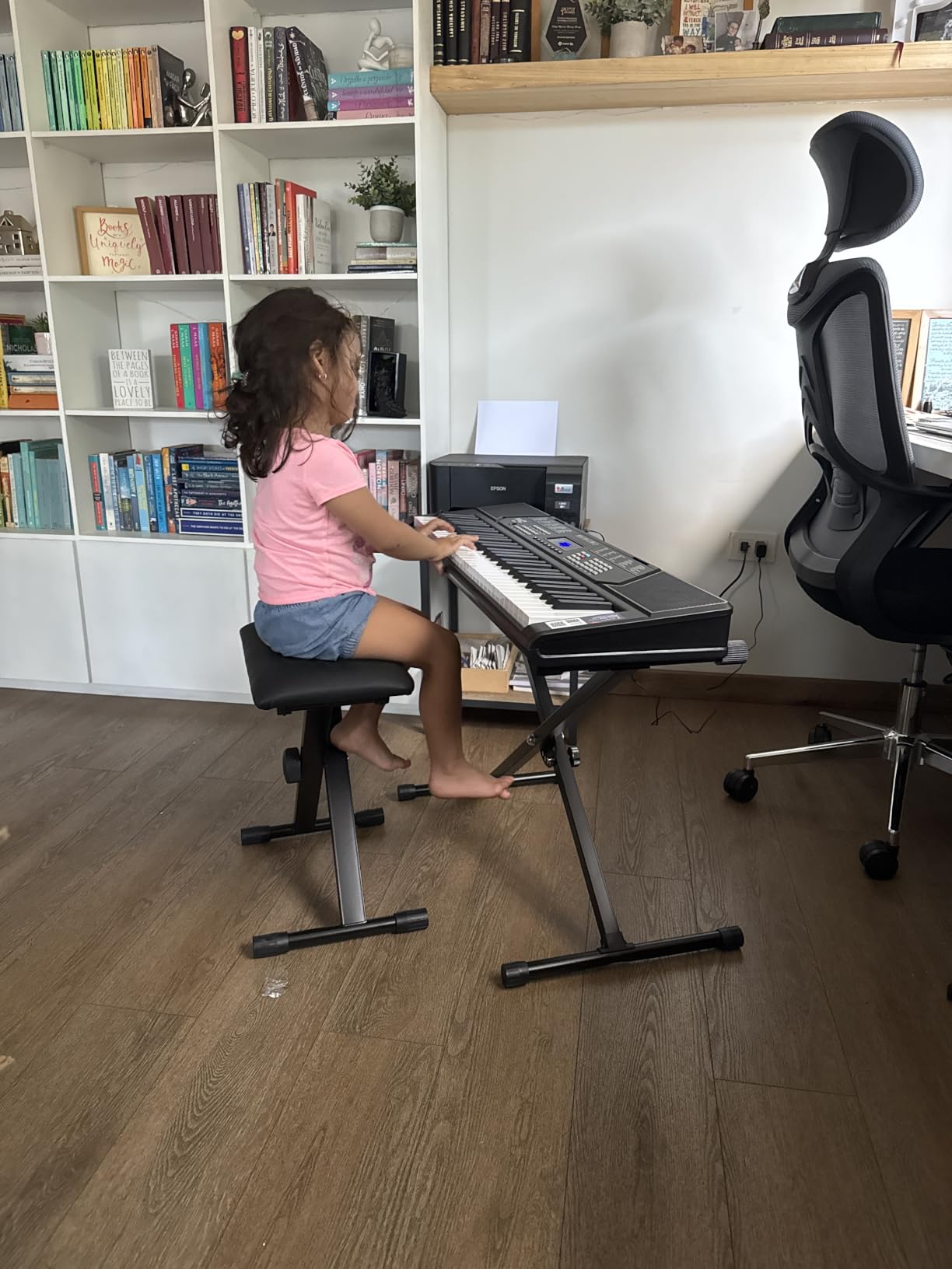 A young child playing a RockJam 61-key keyboard on its X-stand, sitting on the padded bench that came with the kit