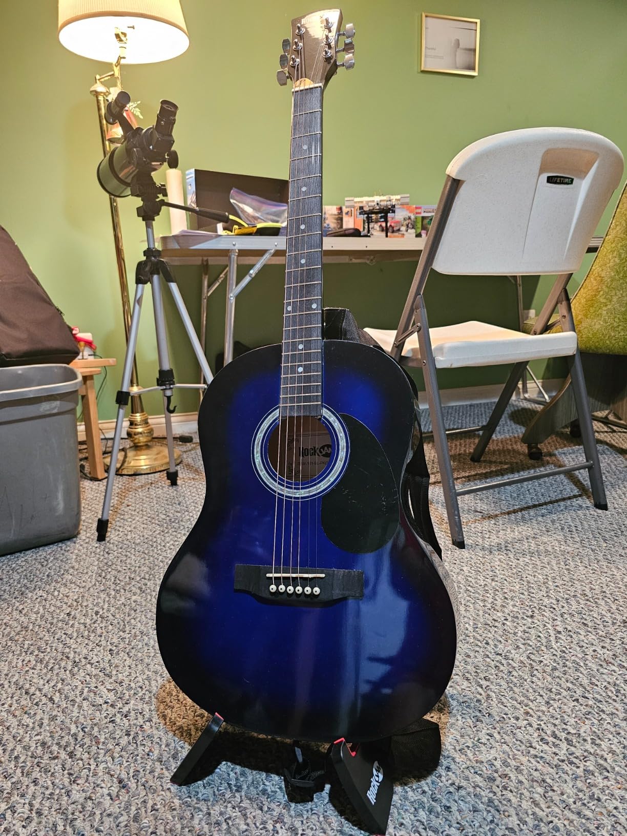 RockJam blue burst acoustic guitar standing on its folding RockJam stand in a living room with the gig bag behind it