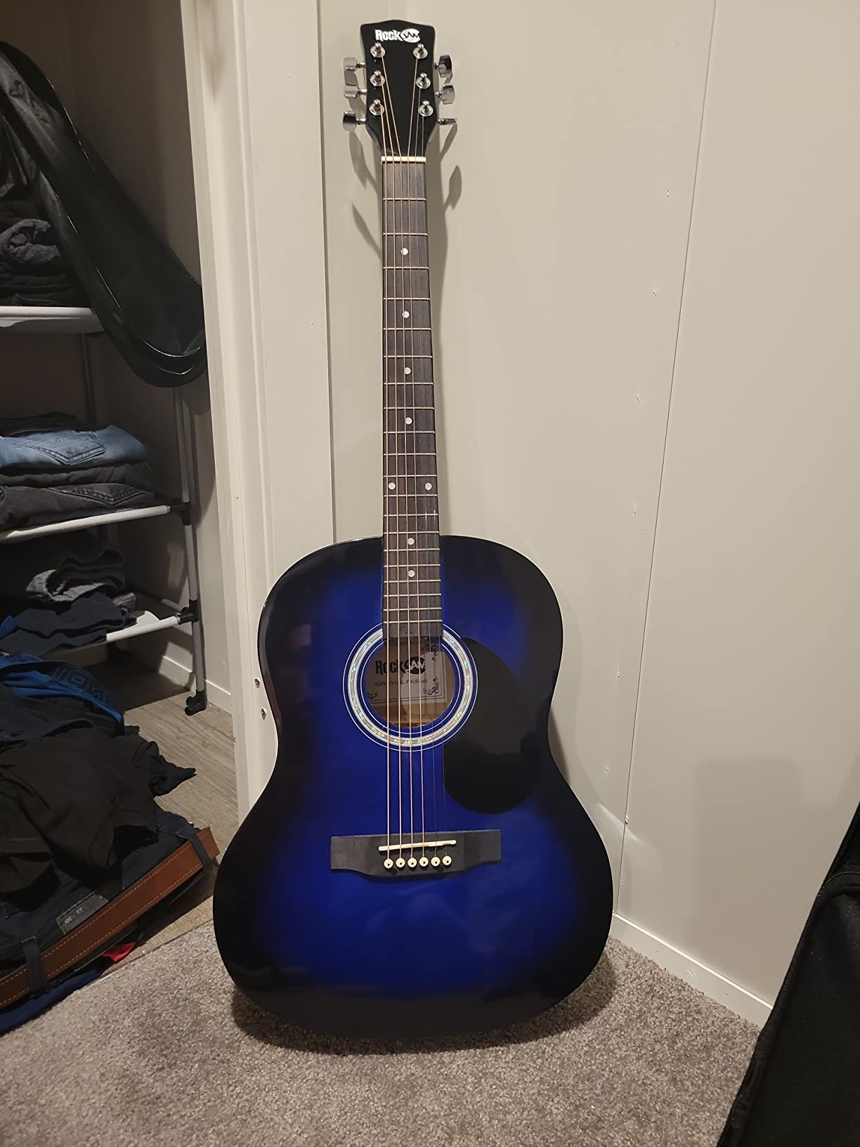 RockJam blue burst steel string acoustic guitar propped against a bedroom wall next to its gig bag